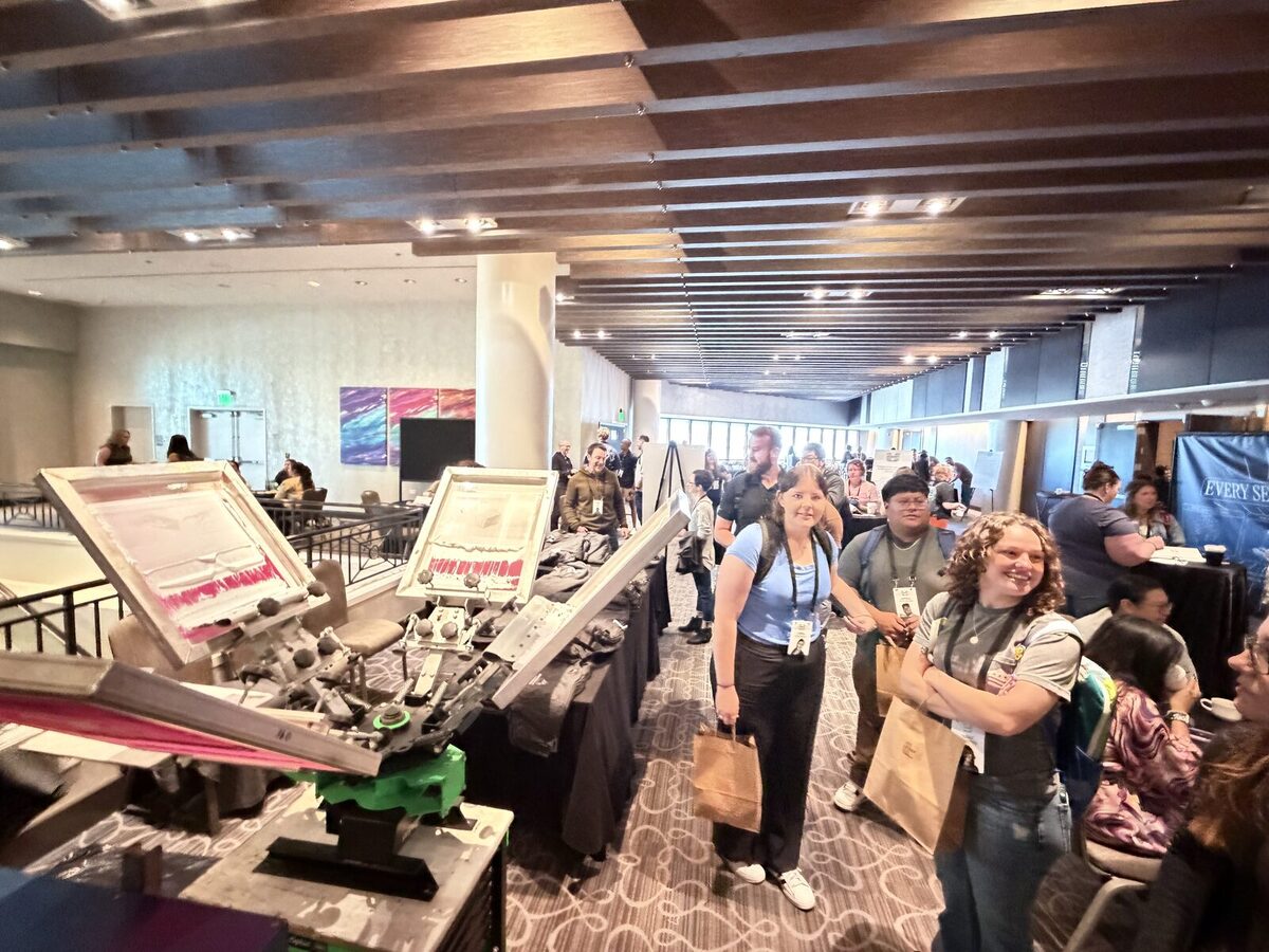 Guests with tote bags walking past screen printing presses set up in a hotel conference foyer