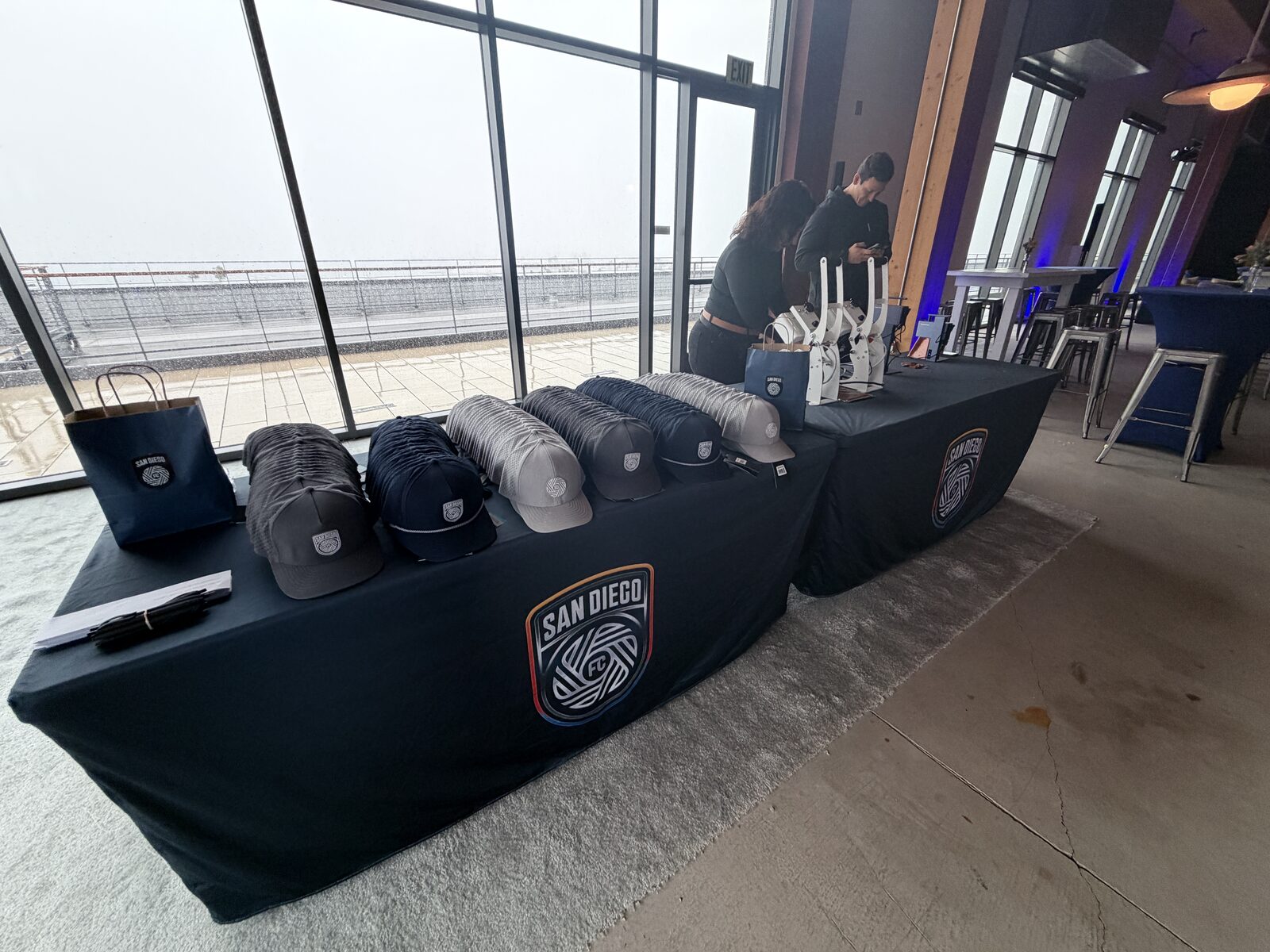 Two long merch tables with grey and navy caps arranged in rows and heat presses at the far end