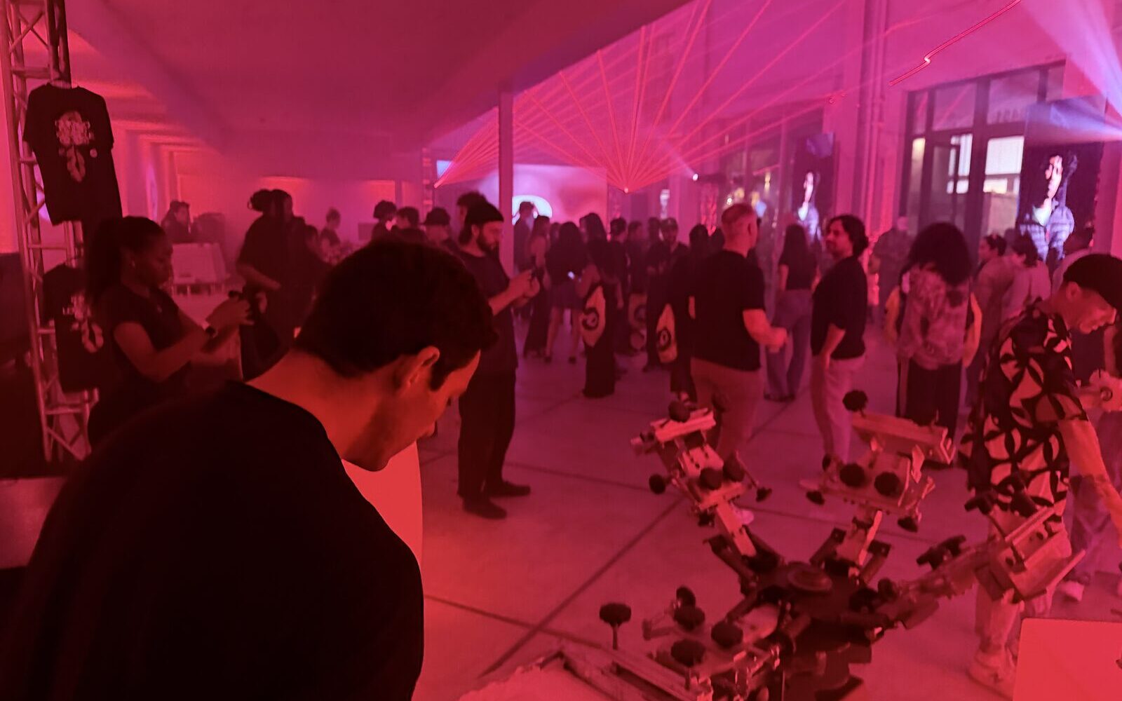 A dim event floor lit pink by lasers with a crowd moving past printing equipment set up in the open