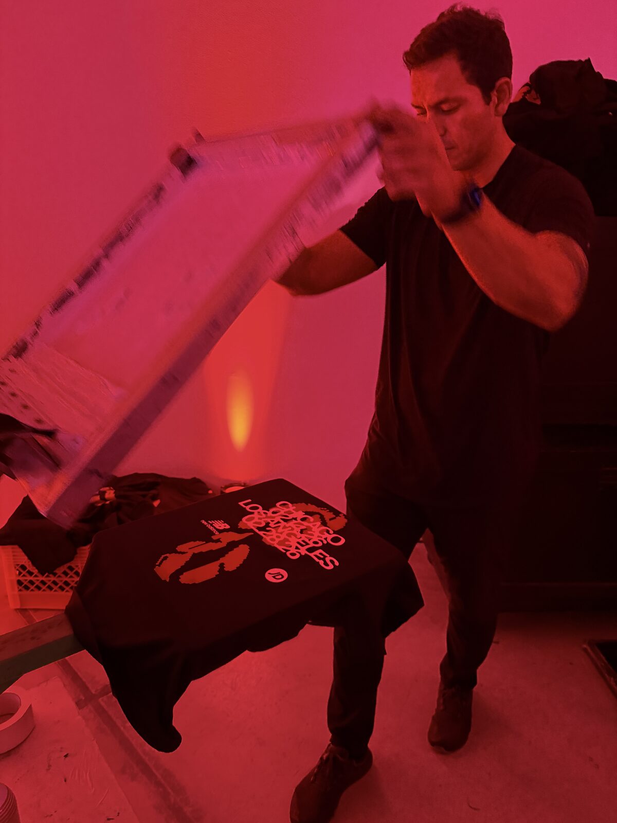 A printer lifting a screen off a black shirt under red event lighting, with a crate of blanks beside the press
