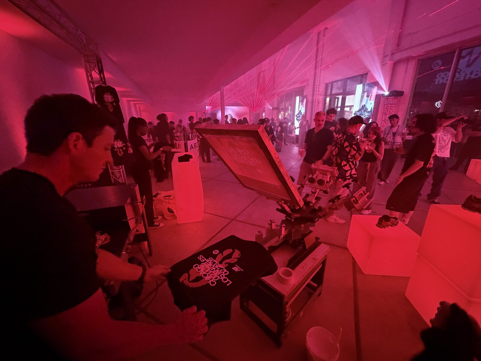 A printer pulling a freshly printed black shirt at an evening party while guests stand around under pink laser lighting