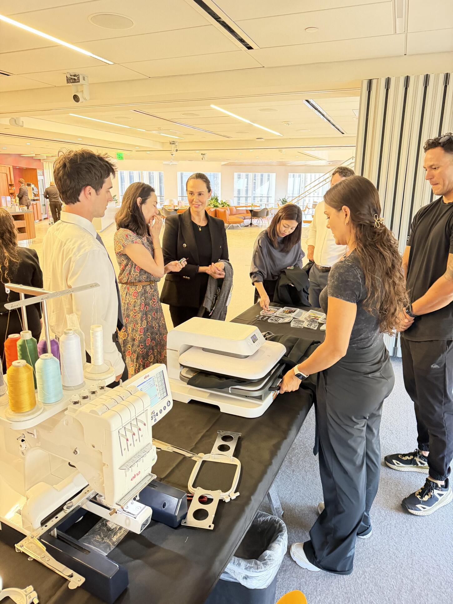 Guests gathered around an indoor personalization counter with a heat press and a multi-needle embroidery machine while a crew member works