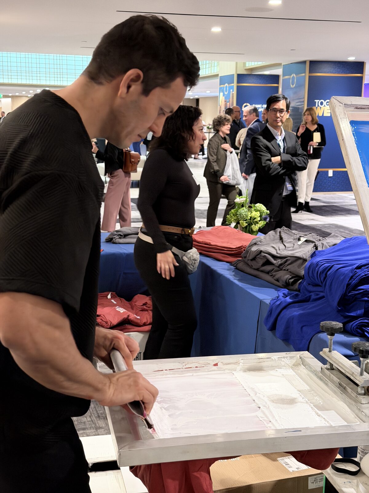 Two guests leaning in to watch a printer pull ink across a screen at an indoor event, with folded shirts stacked on tables behind them