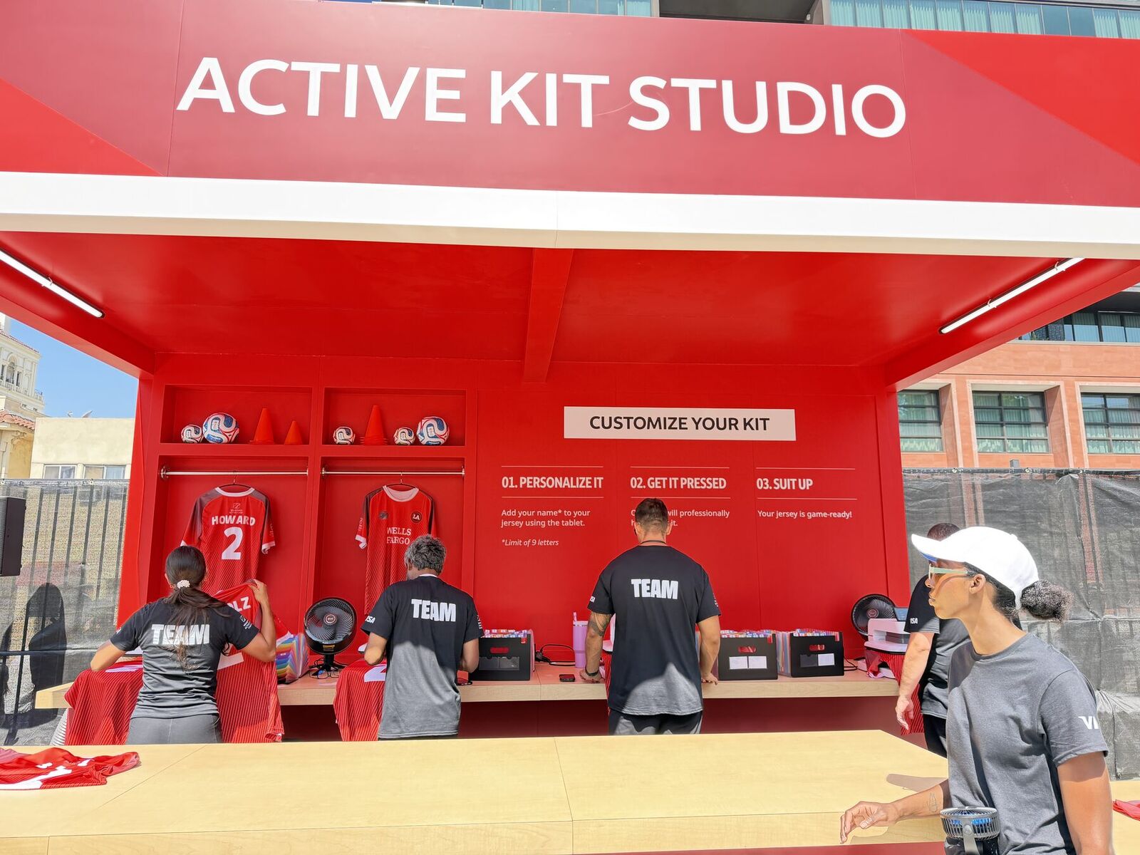 A red outdoor kit studio with jerseys hanging in locker bays, a printed three-step sign reading personalize it, get it pressed, suit up, and a crew in black TEAM shirts working the counter
