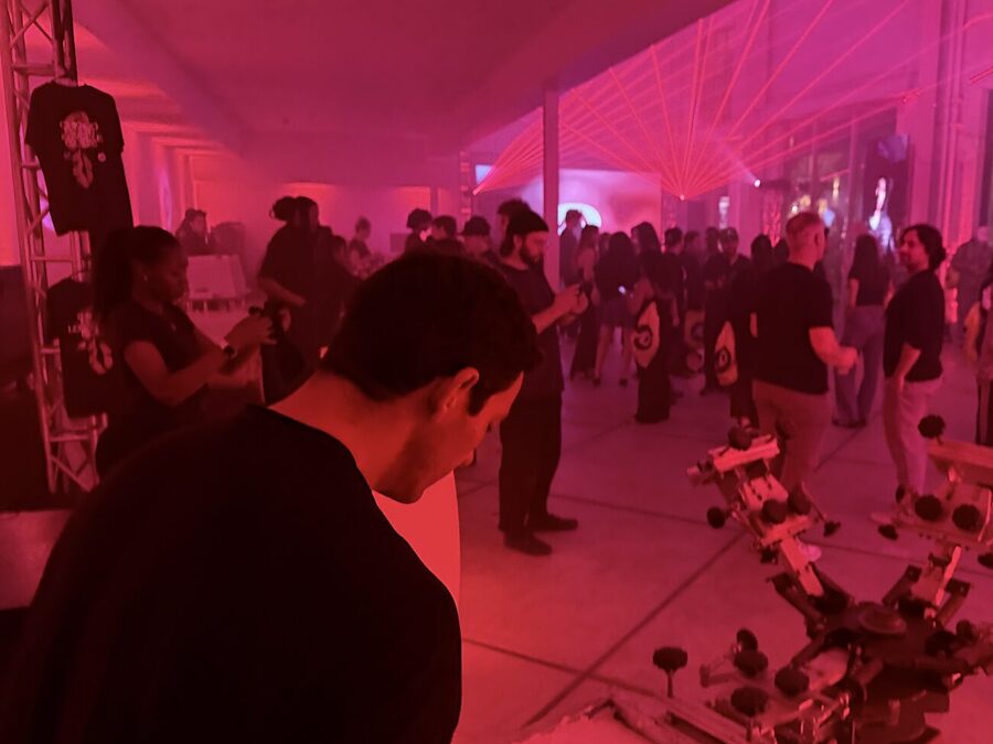 A dim party floor lit red with guests standing among screen printing presses and a printed tee hanging on truss